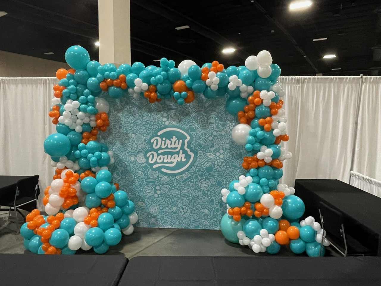 A Dirty Dough branded backdrop with a modern organic balloon arrangement in teal, orange, and white — an eye‑catching setup ideal for high‑visibility corporate booths.